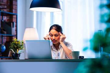 Indian businesswoman feels pressure and headache while working on laptop at modern office desk