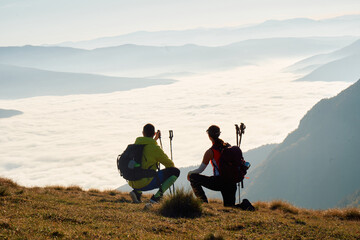 Couple hikers trek up a rocky mountain trail, enjoying a scenic view above the clouds, with majestic peaks in the background.