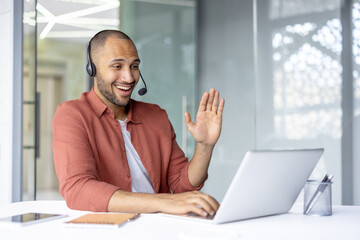 Support service worker with headset smiling, talking and consulting customers online remotely. Man...