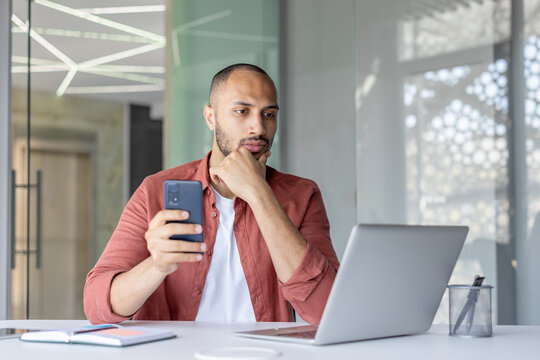 A thoughtful man in a modern office setting, using his smartphone and laptop while considering information and making decisions.