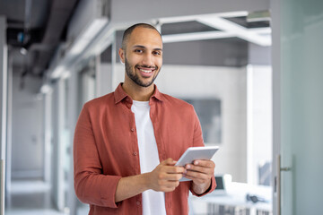 A smiling, bald man in a red shirt holds a tablet in an office setting with a blurred background.