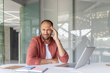 A man at a desk looks stressed while working on his laptop in a modern office environment.
