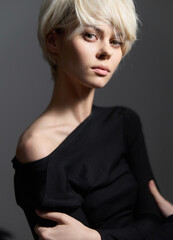 Elegant young woman with short blonde hair, wearing a black off-shoulder top, gazes confidently at...