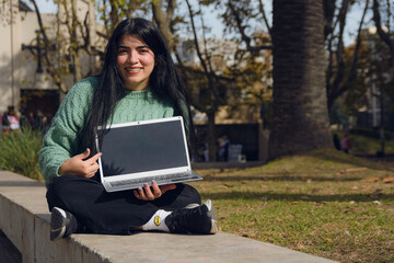 Young Venezuelan woman sitting outdoors with her laptop showing the screen and looking at camera