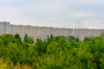 Row of identical gray apartment blocks behind lush green trees under cloudy sky, post-communist housing estate concept