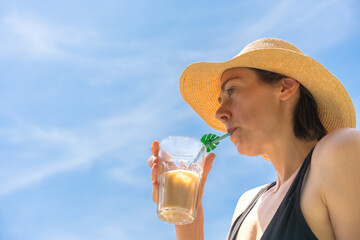 Woman drinking iced coffee outdoors under blue sky on sunny day wearing straw hat, leisure refreshment and summer hydration. large copy space