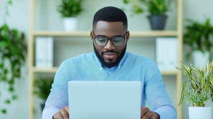 Portrait of a young African man working intently on his laptop computer in a bright modern office environment with minimalist decor and greenery accents