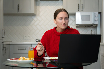 Young woman enjoying a healthy breakfast while working remotely on her laptop in a stylish kitchen,...