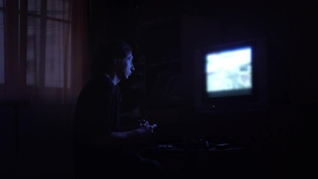 An intensely absorbed and atmospheric mood is captured as a man plays a video game in a dark room, his face illuminated by the blue glow of an older television screen