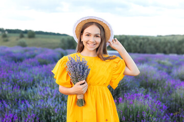 Beautiful young girl in straw boater hat and a yellow dress collects lavender on lavender field....