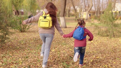 Cheerful mother and child run with backpacks their shoulders autumn park, happy family life, run...