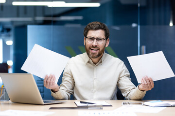 Portrait of angry young male manager sitting at a desk in the office, holding documents in his hands and shouting at the camera in frustration