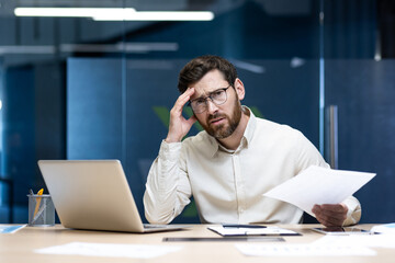 Portrait of a tired and upset young businessman sitting in the office at his desk, working on a...