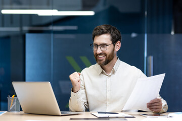 A happy young businessman is sitting at a desk in the office, holding a document, looking at the...