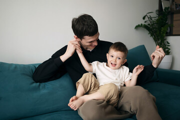 Happy father and cheerful son playing together and smiling on a teal sofa in cozy modern home...