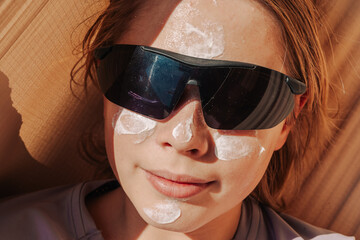 Teen applying sunscreen on face, arm, and leg under direct sun. Summer skincare, blue sky, beach background, and beige hammock. Sun protection routine, outdoor wellness