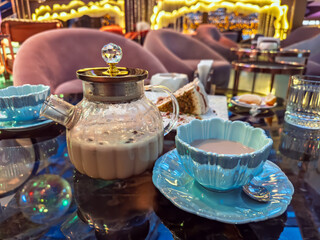 A chic tea time setup with a glass teapot, elegant blue cup, and dessert in elegant cafe.Elegant glass teapot and a stylish blue cup of milk masala tea served in a modern, upscale restaurant lounge