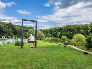 Girl in a hoodie relaxing on a large modern swing at a summer resort overlooking a pool and green forest.Summer leisure and recreation concept. A person rests on a swing in a beautiful resort area