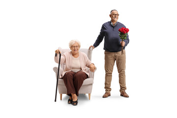 Mature man holding red roses and standing next to a senior woman seated in an armchair