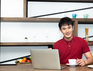 Young Smiling handsome wearing red t-shirt meeting online and drinking a cup of coffee and
work from home. Communicating online, writing emails, distantly working or studying on computer.
