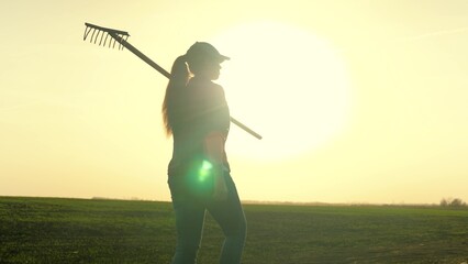 A woman agronomist walks across a field of land with a rake on her shoulders to work, a farmer at...