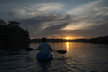 A man paddles a kayak on a tropical island with a sunset in the background. A man enjoys the sunset while playing kayak