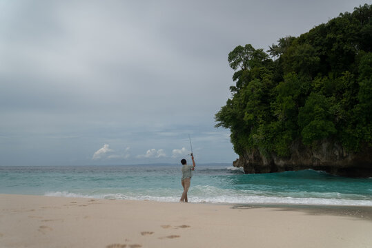 A man fishing off the coast of a tropical island. An angler uses a rod to pull a fish from the choppy shoreline and turquoise water