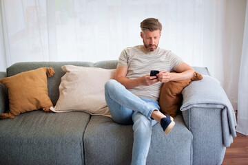 Middle aged man sitting on the couch at home and using his cell phone