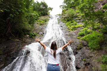 Back view of young asian woman walk in front of waterfall with her hands raised.Female tourist walks in Thailand ,tropical nature in sarika waterfall at nakhon nayok province, Thailand