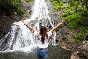 Back view of young asian woman walk in front of waterfall with her hands raised.Female tourist walks in Thailand ,tropical nature in sarika waterfall at nakhon nayok province, Thailand