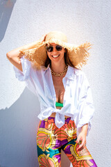 Confident woman wearing sun hat and sunnies agains standing at white wall in the sunshine
