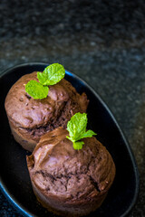 Two chocolate mint flavoured muffins, decorated with mint leaves, in a small, black elongated plate on a shiny stone table top.