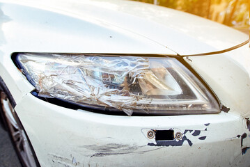 Close-up view of white car damaged bumper and broken headlight covered with tape, recent crash and...