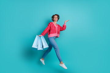 Full length photo of sweet excited lady wear red cardigan holding bargains jumping emtpy space isolated turquoise color background