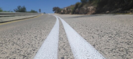 A low-angle view of a road with sharp focus on the double white lines, leading uphill under a clear...