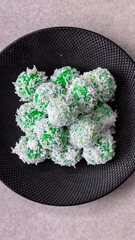 Traditional Indonesian klepon, green sticky rice balls with palm sugar, coated in shredded coconut, served on a black plate.
