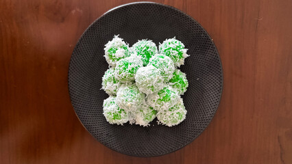 Traditional Indonesian klepon, green sticky rice balls with palm sugar, coated in shredded coconut, served on a black plate over a wooden table.