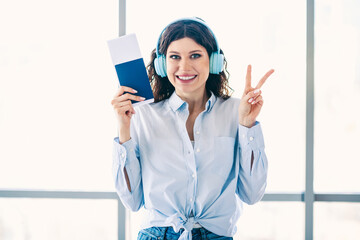Happy young woman with passport and headphones showing a peace sign while at the airport, excited...