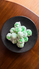 Traditional Indonesian klepon, green sticky rice balls with palm sugar, coated in shredded coconut, served on a black plate over a wooden table.