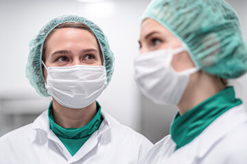 Two healthcare professionals wearing surgical masks and hairnets engage in a conversation in a clinical setting.