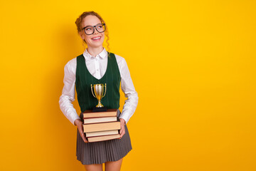Smiling red-haired student in green uniform holding books and a trophy, standing against a yellow...