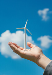 A hand holds a small white wind turbine, with a blue sky and clouds in the background, symbolizing clean energy
