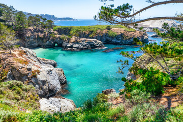 Breathtaking view of China Cove at Point Lobos State Natural Reserve, California. Turquoise waters, rocky cliffs, and vibrant coastal vegetation under a clear summer sky