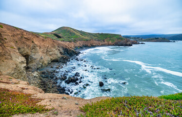Sweeping view from Mori Point in Pacifica, California. Grassy bluff overlooks the Pacific Ocean and distant Pacifica Pier under an overcast sky, capturing the rugged beauty of the coastline
