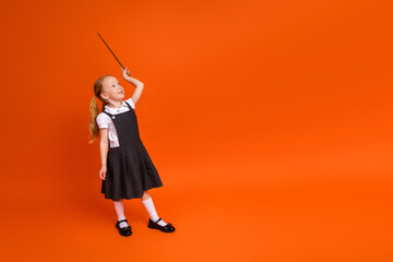 Adorable little girl in a school uniform with ponytails cheerfully raising a stick on an orange...