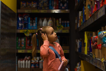 A pretty little girl chooses what to buy in a supermarket, holding her hand near her head, looking...