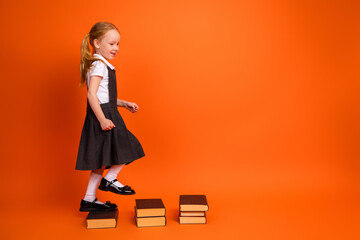 Adorable schoolgirl stepping on stacked books against vibrant orange studio background symbolizing...