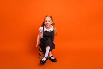 Cheerful young schoolgirl sitting on books against an orange background, showcasing a bright...