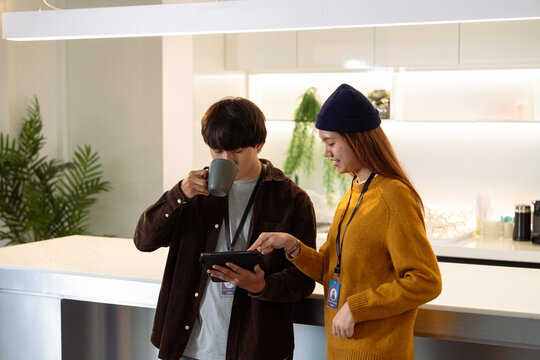 Two young adults engage in conversation while one holds a mug and the other uses a tablet in a modern kitchen setting.