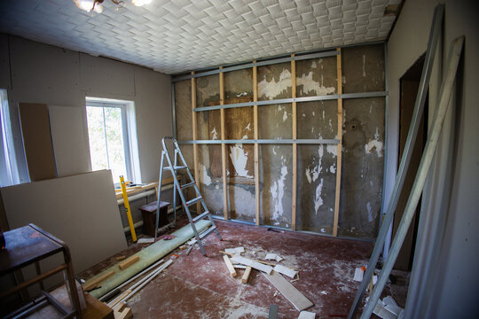 Metal frame attached to the wall with wooden bars and screws, preparing for plasterboard installation in a renovated room with scattered construction materials and a nearby ladder.